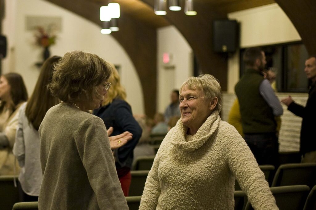 Two women talking and holding hands after a Friday gathering, with others in conversation behind them